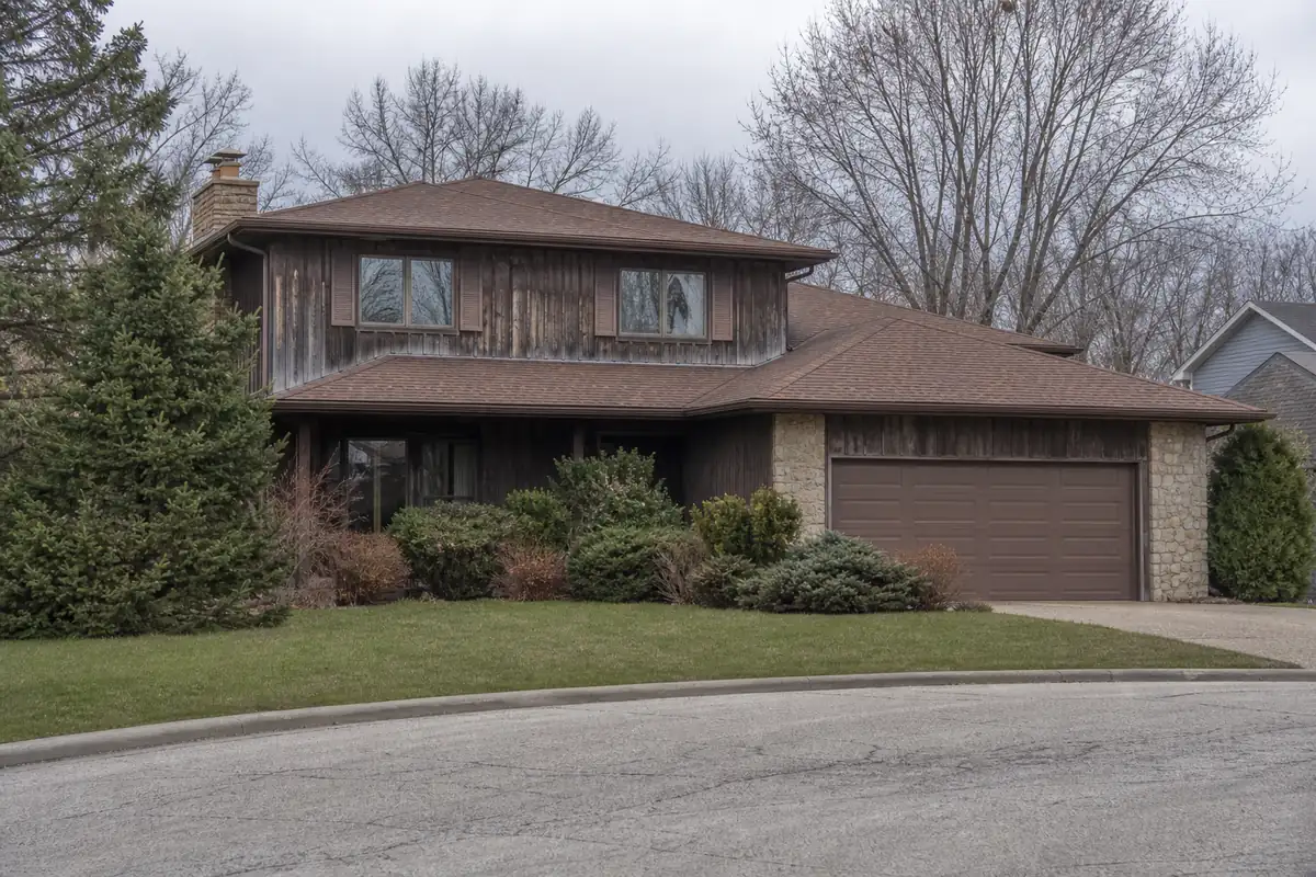 A late-1970s two-story house with cedar siding on a curving subdivision street — we buy houses waukesha — Cudahy, WI